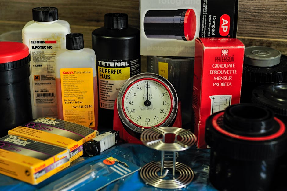 A close-up shot of a collection of various household items and photographic chemicals arranged on a wooden surface, including bottles of Ilford and Kodak photographic chemicals, a black and red stopwa