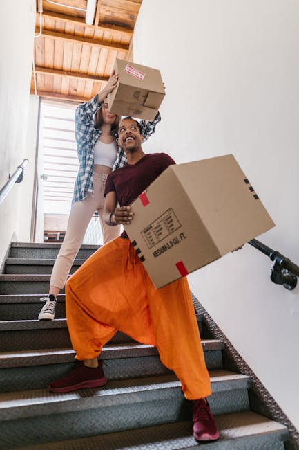 In an indoor home environment, a man with a beard is seen standing on a staircase landing, smiling while holding a cardboard box to assist with a house relocation. The staircase features wooden handra