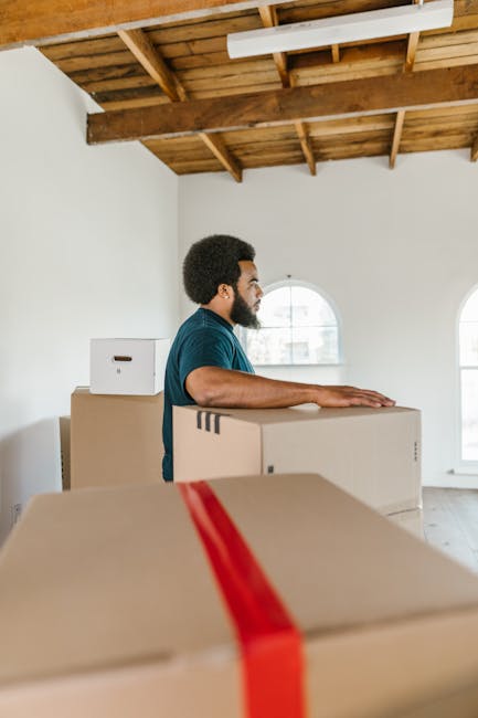 A man with dark curly hair and a beard, dressed in a blue shirt, is seen inside a room with white walls and a wooden ceiling, preparing for a home relocation. He is standing behind a stack of cardboard moving boxes, with one box in the foreground wrapped with red packing tape. The boxes vary in size and are arranged on the floor, indicating the packing and loading process typical of furniture transport and house removals. The room features large arched windows allowing natural light to illuminate the space, and the overall setting suggests the early stages of a local moving service being carried out by Man With a Van Ilford, a company specializing in removals in the Ilford area.