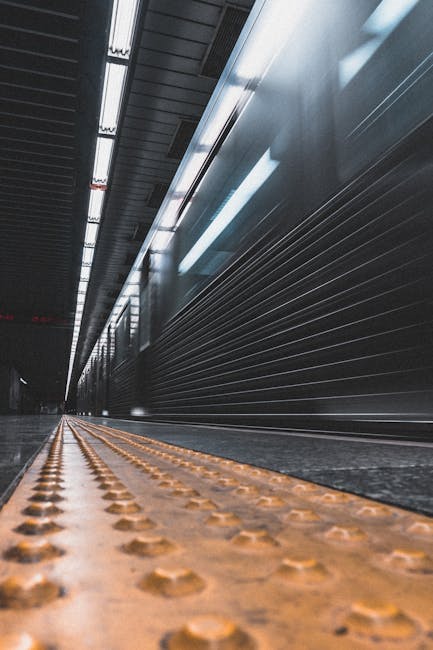 Inside an underground railway station with a low-angle view showing a long platform edge featuring tactile paving strips in orange, adjacent to a dark, metallic wall with horizontal ridges. Overhead, bright fluorescent lights illuminate the corridor, reflecting off the smooth surfaces and creating a contrast between the well-lit area and the darker background. Although not explicitly detailed in the image, this setting aligns with typical urban transport environments where home relocation or furniture transport might intersect with transit logistics. Such a location is representative of the environments where Man With a Van Ilford could coordinate furniture transport and moving services, assisting clients with efficient, local moves within bustling city infrastructure.